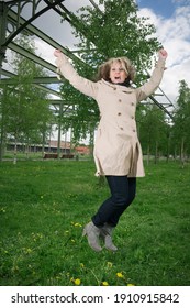 Full length view of a mature woman in a trench coat in a public park with arms up, jumping joyfully.