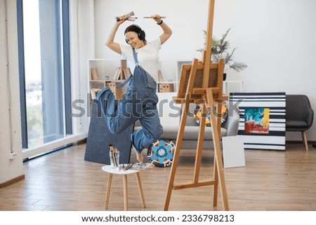 Full length view of passionate female in wireless device jumping in air with paintbrushes in spasious workshop. Ardent music lover imitating rock star while rejoicing concert during artistic process.