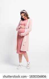 full length view of pregnant woman in cardigan and headband touching belly on white