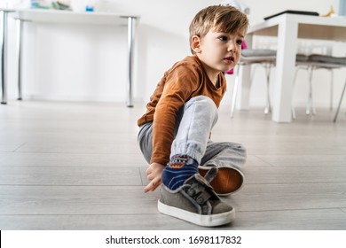 Full Length View Of Small Cute Caucasian Boy Little Child Kid Sitting On The Wooden Or Laminated Vinyl Floor At Home Putting On Or Taking Off The Shoes In Day Alone Front View