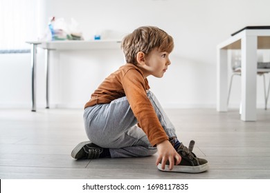 Full Length View Of Small Cute Caucasian Boy Little Child Kid Sitting On The Wooden Or Laminated Vinyl Floor At Home Putting On Or Taking Off The Shoes In Day Alone Side View