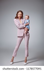 Full Length View Of Stylish Young Mother Holding Adorable Infant Daughter And Looking At Camera On Grey