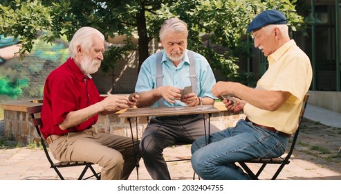 Full Length View Of The Three Caucasian Senior Men Best Friends On Retirement Playing Cards In The Yard At The Table. Outside. Leisure Games And Retirement Concept 