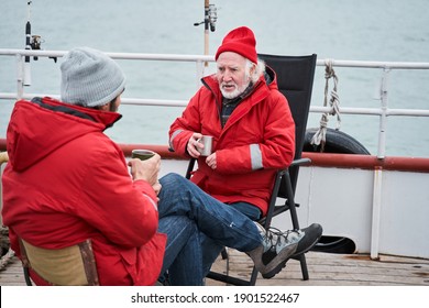 Full length view of the two confident fishermen colleagues discussing something during tea break at the deck. People talking about sea and fish while sitting at the boat. Occupation concept