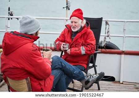 Full length view of the two confident fishermen colleagues discussing something during tea break at the deck. People talking about sea and fish while sitting at the boat. Occupation concept