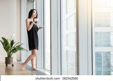 Full Length View Of Young Woman Standing Near Window Enjoying Coffee, Looking At City After Waking Up At Morning, Wearing Black Silk Nightwear, Resting From The Business, Waiting For Good Weather 