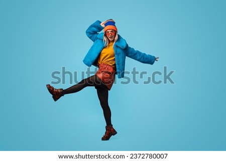 Full length weird young lady in colorful stylish outfit lifting leg and touching hat while dancing against blue background