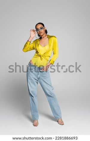 full length of well dressed african american woman in blue sunglasses posing with hand in pocket on grey