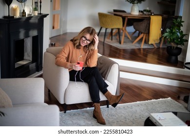 Full length of a woman sitting at home and using earphones and smartphone while having video call.  - Shutterstock ID 2255810423