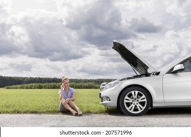 Full length of worried businesswoman sitting by broken down car at countryside