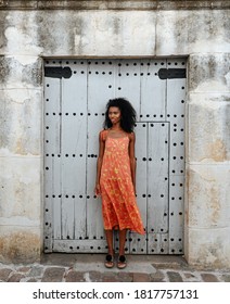 Full Length Of Young African American Female In Colorful Sundress Standing Against Shabby Door Of Old Stone Building On City Street