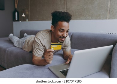 Full Length Young African American Man In T-shirt Lay Down On Sofa Indoors Apartment Use Laptop Pc Computer Credit Bank Card Shopping Online Order Delivery Booking Tour Resting On Weekends Stay Home