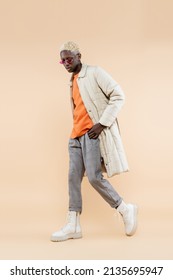 full length of young african american man in stylish sunglasses and coat posing with hand in pocket on beige