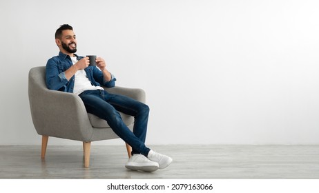 Full length of young Arab man drinking hot coffee in armchair against white studio wall, banner with free space. Peaceful middle Eastern guy having relaxing day, chilling on lazy morning - Shutterstock ID 2079163066