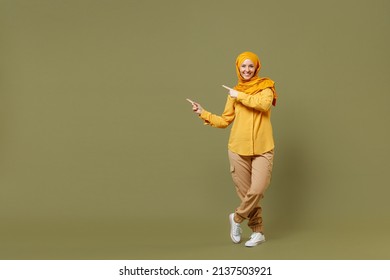 Full length young arabian asian muslim woman in abaya hijab yellow clothes point index finger aside on workspace isolated on olive green background. People uae middle eastern islam religious concept