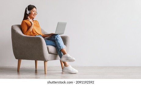 Full length of young Asian female in headphones using laptop for online communication, video chatting, waving at webcam, sitting in armchair against white studio wall, banner design with copy space