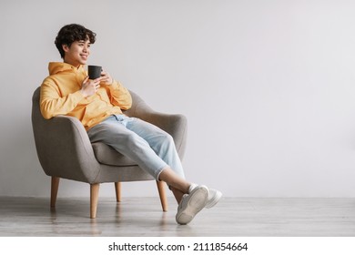 Full length of young Asian man drinking hot coffee in armchair against white studio wall, empty space. Peaceful millennial guy having relaxing day, chilling on lazy morning with warm beverage