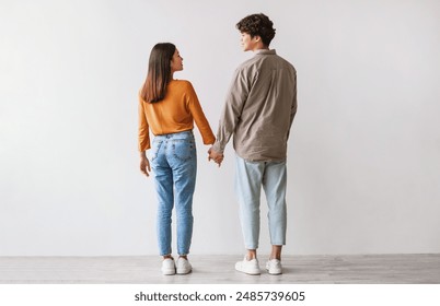 Full length of young Asian man and woman standing with their backs to camera, looking at each other and holding hands against white studio wall. Millennial couple expressing affection - Powered by Shutterstock