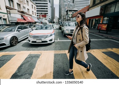 Full Length Of Young Asian Office Lady Walking On Zebra Cross Going To Work. Girl Backpacker Holding Cell Phone Searching Online Map In Busy City Urban. Cars Vehicle Waiting Stop Traffic Light