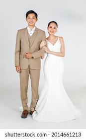 Full length of young attractive Asian couple, bride and groom, woman wearing white wedding dress. Man wearing beige suit, standing together arm in arm. Concept for pre wedding photography.