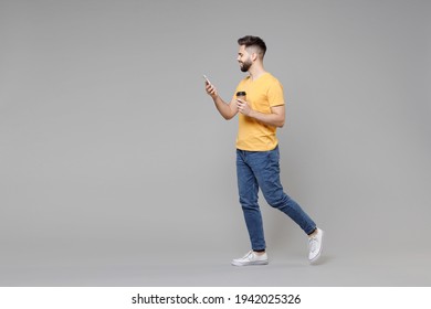 Full length of young bearded attractive smiling student man 20s in casual yellow basic t-shirt drinking coffee holding paper cup of tea, mobile cell phone isolated on grey background studio portrait. - Powered by Shutterstock