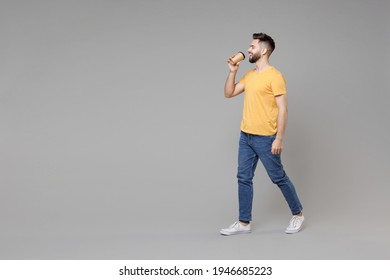 Full length of young bearded attractive smiling student man 20s in casual yellow basic t-shirt walking drinking coffee holding paper cup of tea isolated on grey color background studio portrait.