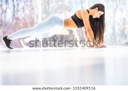 Full length of young beautiful woman in sportswear doing plank while standing in front of window at gym