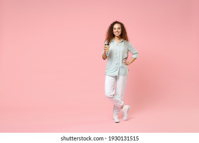 Full length young black african fun happy smiling stylish curly student woman 20s in blue shirt holding paper cup of coffe arm akimbo on waist isolated on pastel pink color background studio portrait