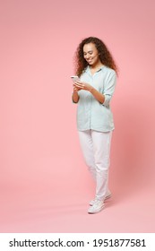 Full length young black african fun happy smiling curly student woman 20s in blue shirt holding mobile cell phone chatting browsing internet isolated on pastel pink color background studio portrait