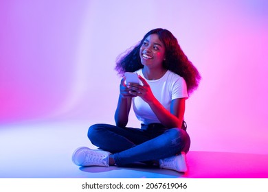 Full length of young black lady sitting cross legged with smartphone, browsing social media, messaging to friend in neon light. African American woman working or studying on mobile device, copy space - Shutterstock ID 2067619046