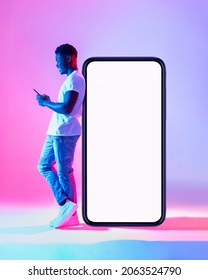 Full length of young black man using mobile phone while leaning on huge smartphone with empty white screen in neon light. Mockup for cool new app or website, space for ad - Shutterstock ID 2063524790