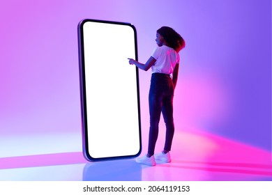 Full length of young black woman touching screen of giant smartphone in neon light, mockup for new mobile app or website design. African American lady interacting with big cellphone, space for ad - Shutterstock ID 2064119153