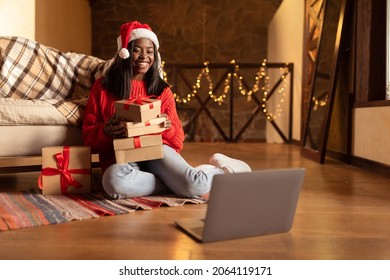 Full length of young black woman holding Xmas gift boxes in front of laptop, speaking to friends online, congratulating family with Christmas holidays, sitting on floor at home, copy space - Powered by Shutterstock