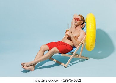 Full Length Young Blond Man In Red Shorts Swimsuit Sit In Deckchair Near Hotel Pool Hold Inflatable Rubber Ring Drink Beer Isolated On Pastel Blue Background Summer Vacation Sea Rest Sun Tan Concept
