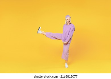 Full Length Of Young Blonde Caucasian Woman 20s Bob Haircut In Casual Basic Purple Suit Beanie Hat Dancing Raised Leg Up Spreading Hands Fooling Around Isolated On Yellow Background Studio Portrait