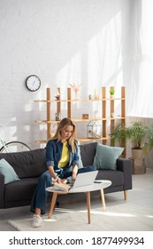 Full Length Of Young Blonde Woman Using Laptop While Sitting On Couch Near Coffee Table At Home