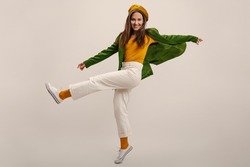 Full Length Young Caucasian Girl Moves Vigorously Waving Legs On White Background In Studio. Brunette Wears Yellow Beret, Blouse, Green Jacket And Pants. Good Mood, Fashion Trends