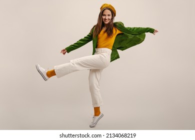 Full length young caucasian girl moves vigorously waving legs on white background in studio. Brunette wears yellow beret, blouse, green jacket and pants. Good mood, fashion trends - Shutterstock ID 2134068219
