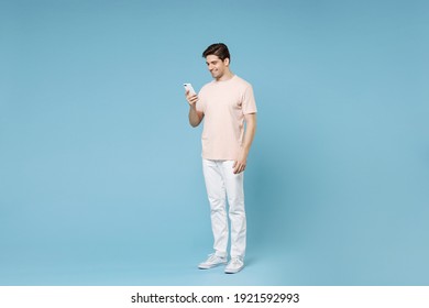 Full Length Of Young Caucasian Happy Smiling Student Man 20s Wearing Beige T-shirt White Pants Holding Mobile Cell Phone Chatting With Girlfriend Isolated On Blue Color Background Studio Portrait