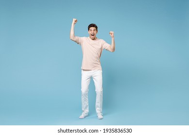 Full length of young caucasian happy excited successful student man 20s in beige t-shirt white pants doing winner gesture clenching fists look camera isolated on blue color background studio portrait