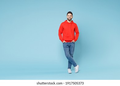 Full Length Young Caucasian Smiling Bearded Handsome Student Man 20s Wearing Casual Red Orange Hoodie Standing Akimbo Arms On Waist Isolated On Blue Background Studio Portrait People Lifestyle Concept