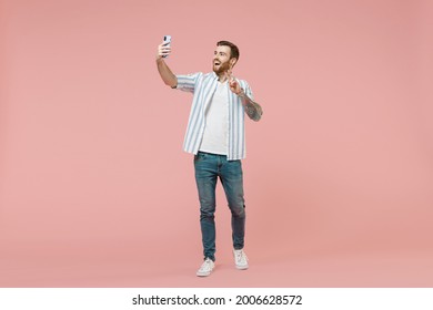 Full length young caucasian unshaven man in blue striped shirt doing selfie shot on mobile phone post photo on social network waving hand isolated on pastel pink background. People lifestyle concept - Powered by Shutterstock