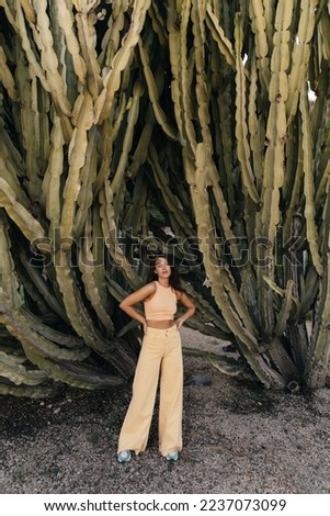 In full length of young caucasian woman looking at camera stands near big cactuses in background. Brunette wears top and jeans. Tourism, vacation concept.