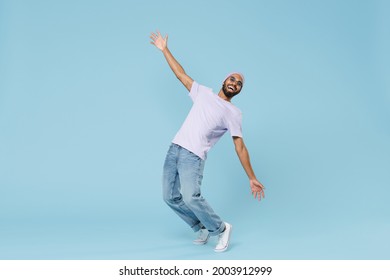 Full length young cool smiling happy funny unshaven black african man 20s wearing violet t-shirt hat glasses standing on toes leaning back dancing isolated on pastel blue background studio portrait