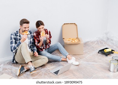 Full Length Of Young Couple Eating Pizza While Sitting Near Laptop On Floor At Home