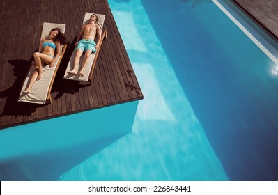 Full Length Of A Young Couple Resting On Sun Loungers By Swimming Pool