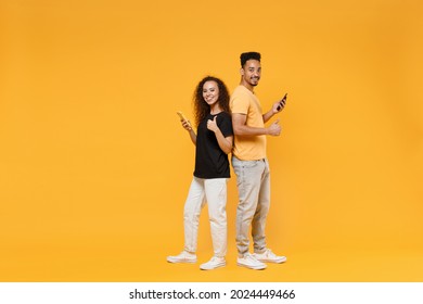 Full Length Young Couple Two Friends Fun Together African Happy Woman Man In Black T-shirt Show Hold Mobile Phone Stand Back To Back Show Thumb Up Gesture Isolated On Yellow Background Studio Portrait