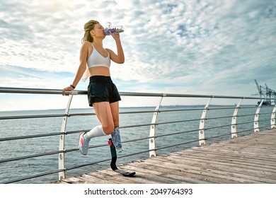 Full length of young disabled athlete woman with prosthetic leg standing on the bridge and drinking water from a bottle. Copy space. - Powered by Shutterstock