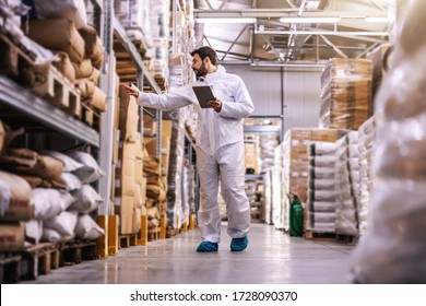 Full Length Of Young Employee In Sterile Protective Uniform Checking On Products While Standing In Warehouse Full Of Food Products In Boxes.