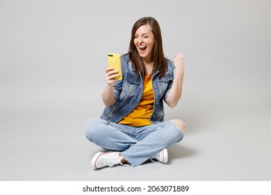 Full Length Young Excited Caucasian Woman 20s In Casual Denim Jacket Yellow T-shirt Use Mobile Cell Phone Browsing Sitting Cross-legged Do Winner Gesture Isolated On Grey Background Studio Portrait.